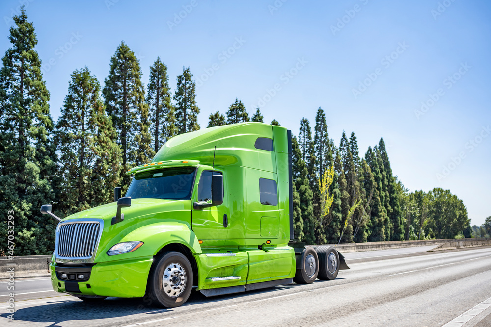 Light green big rig professional semi truck tractor driving on the wide multiline highway road to warehouse for pick up loaded semi trailer for the next freight