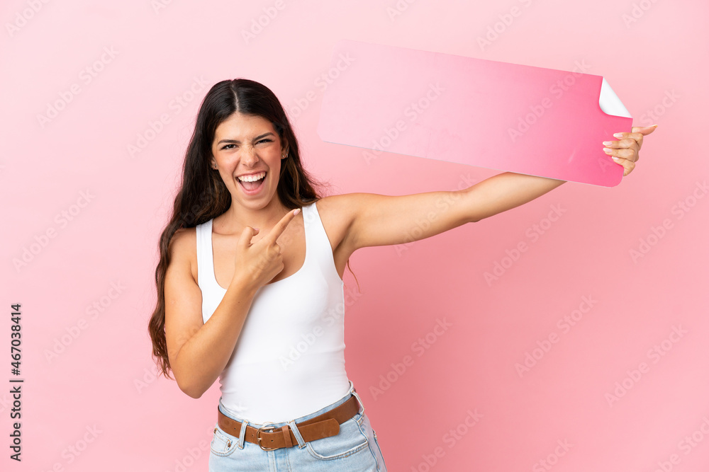 Young caucasian woman isolated on pink background holding an empty placard and pointing it Young caucasian woman isolated on pink background holding an empty placard and pointing it