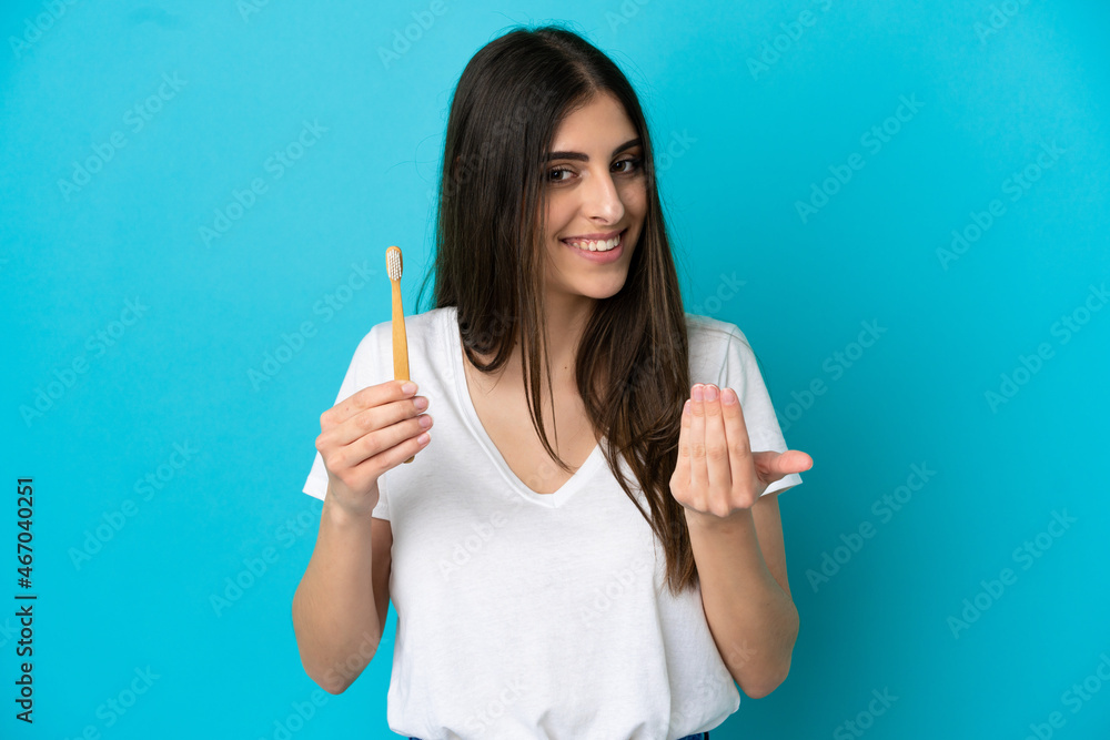Young caucasian woman brushing teeth isolated on blue background inviting to come with hand. Happy that you came Young caucasian woman brushing teeth isolated on blue background inviting to come with hand. Happy that you came