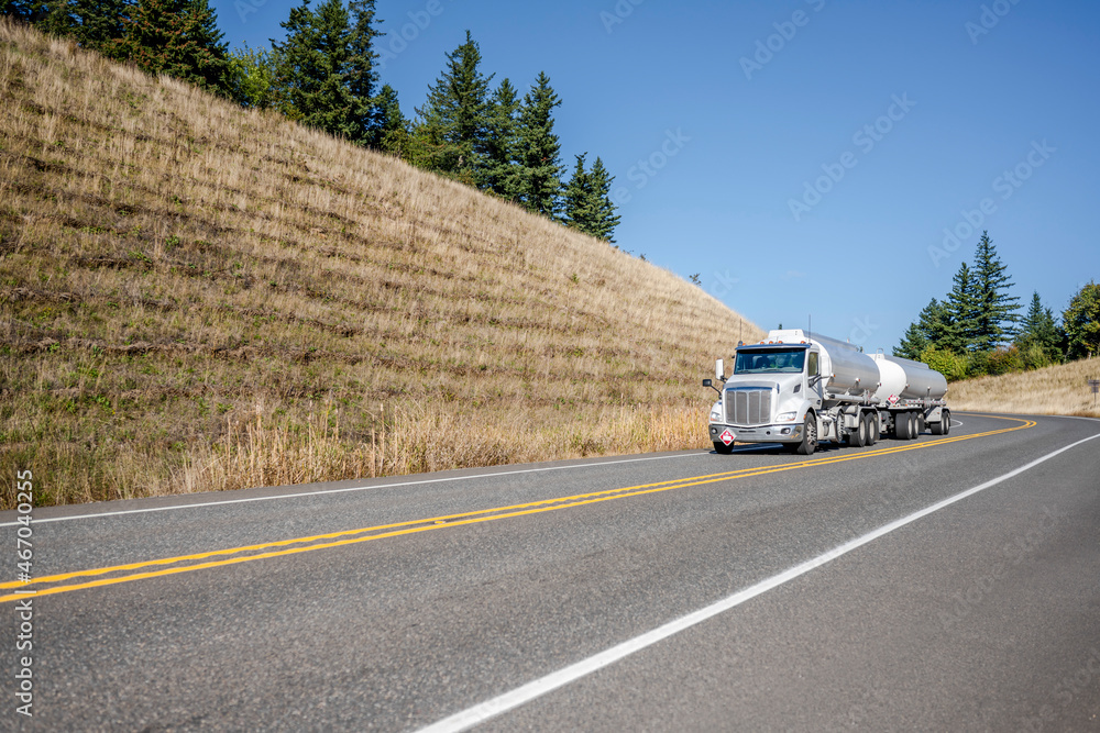 White day cab big rig semi truck transporting hazardous liquid cargo in two tank semi trailers running on the narrow road between hills