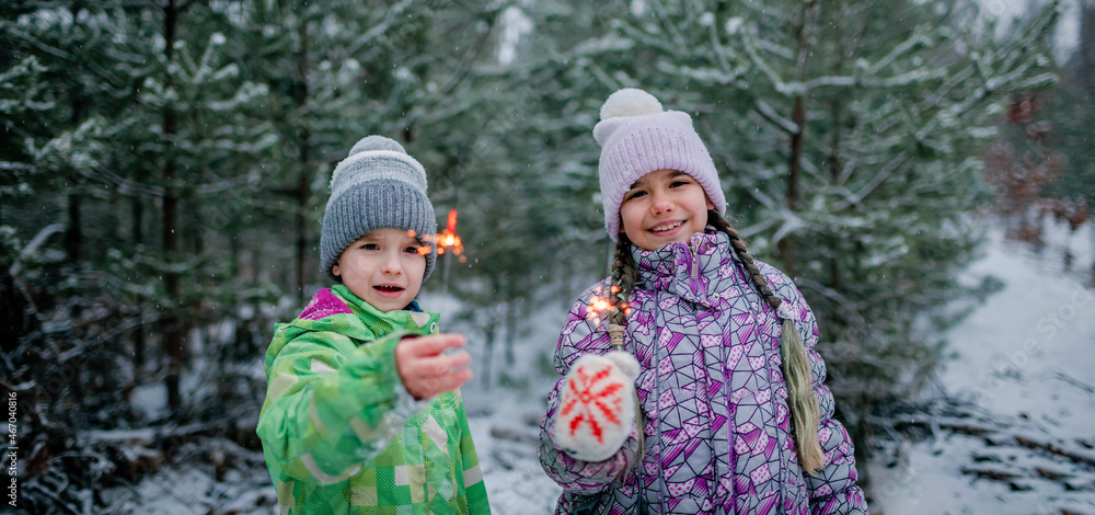 Happy kids with burning sparklers during walking in forest on snowing day, outdoor family Christmas celebration, excited family in warm clothes