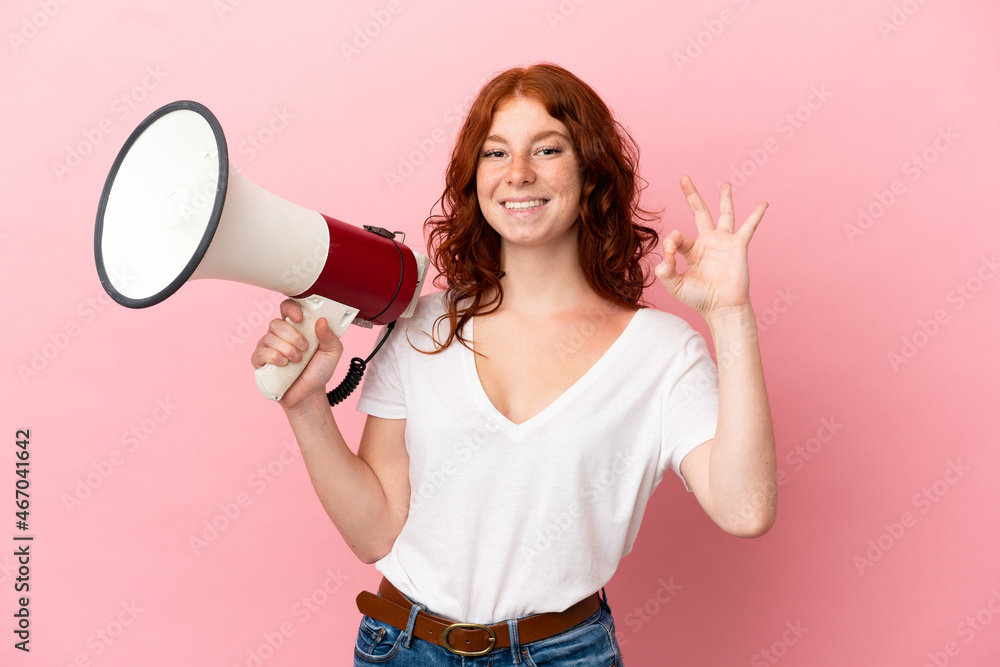 Teenager reddish woman isolated on pink background holding a megaphone and showing ok sign with fingers Teenager reddish woman isolated on pink background holding a megaphone and showing ok sign with fingers