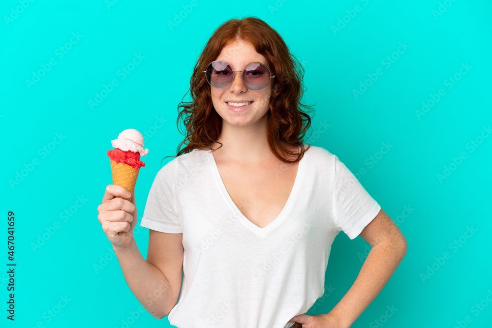 Teenager reddish woman with a cornet ice cream isolated on blue background posing with arms at hip and smiling Teenager reddish woman with a cornet ice cream isolated on blue background posing with arms at hip and smiling