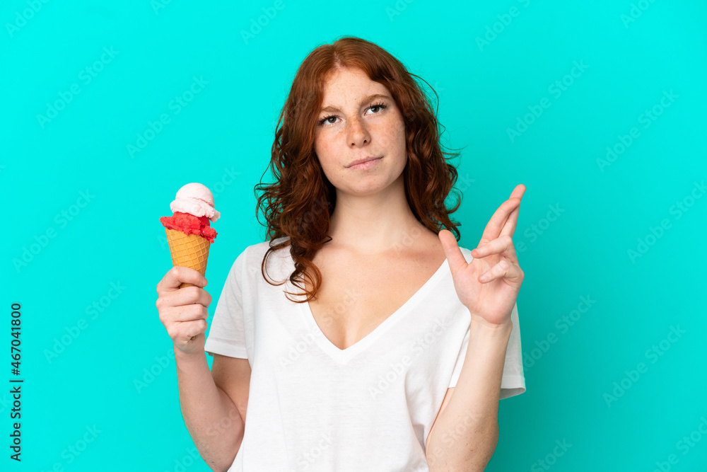 Teenager reddish woman with a cornet ice cream isolated on blue background with fingers crossing and wishing the best Teenager reddish woman with a cornet ice cream isolated on blue background with fingers crossing and wishing the best