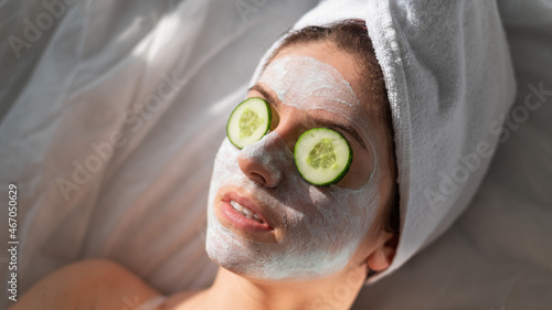 A woman with a towel on her hair and in a clay face mask and cucumbers in front of her eyes lies on the sheet