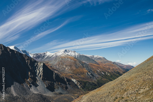 Colorful alpine landscape with great mountain in autumn colors with snow on peak in sunshine under cirrus clouds in blue sky. Picturesque autumn scenery with sunlit rocks and snow-covered mountain top