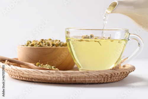 Chamomile tea in a teapot pouring into cup glass with dried chamomile flowers in a bowl on white background, Healthy herbal tea
