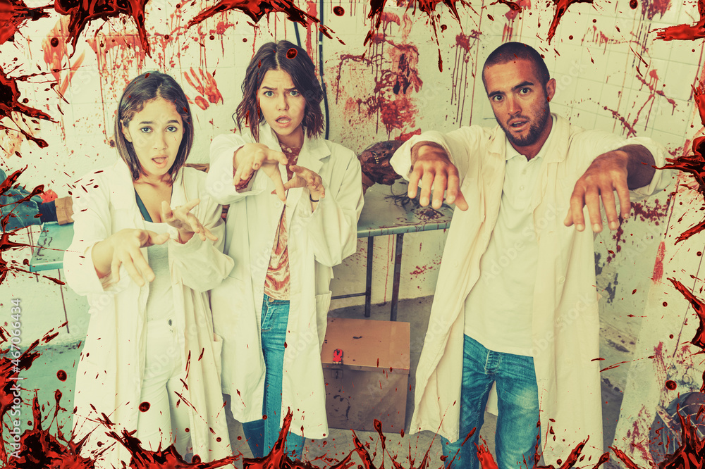 Group of cheerful young adults posing as zombies in stylized escape room with traces of blood