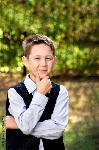Portrait of a schoolboy looking thoughtfully to the side, against a blurry background of green trees.