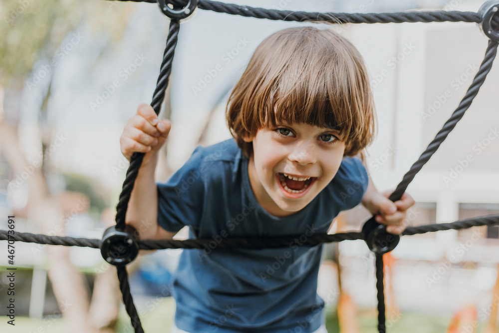 pretty little brunette boy in a blue T-shirt is climbing a rope web. child is engaged in outdoor sports. healthy active kid. lifestyle. space for text. High quality photo pretty little brunette boy in a blue T-shirt is climbing a rope web. child is engaged in outdoor sports. healthy active kid. lifestyle. space for text. High quality photo