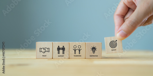 Business goals achievement concept with collaboration and teamwork. Businessman holds the wooden cube with symbols ; goals, trust, support, teamwork and inspiration on grey background and copy space.