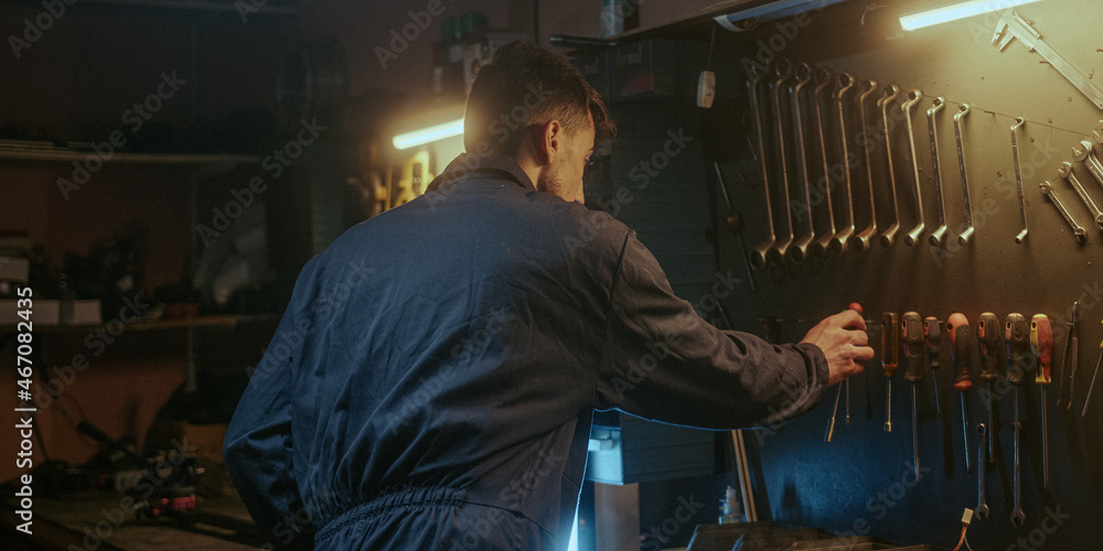 Young determined blacksmith in blue overalls tightens screw with screwdriver and puts his tool in place