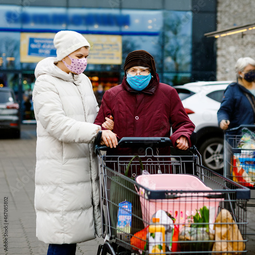 Senior woman and social worker with medical mask due pandemic coronavirus disease. Daughter or granddaughter help grandmother with shopping in supermarket, push cart trolley with foods, outdoors