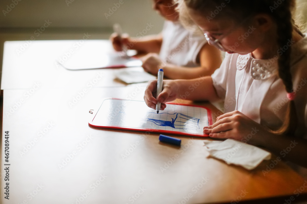 Cute caucasian girl child blonde with pigtails writes with a marker on a tablet, classes at school and independent work of children, light real class at school, soft focus and warm toning Cute caucasian girl child blonde with pigtails writes with a marker on a tablet, classes at school and independent work of children, light real class at school, soft focus and warm toning