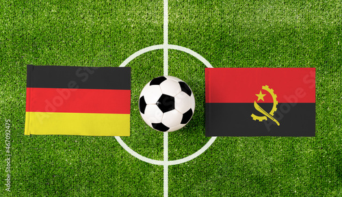 Top view soccer ball with Germany vs. Angola flags match on green football field.