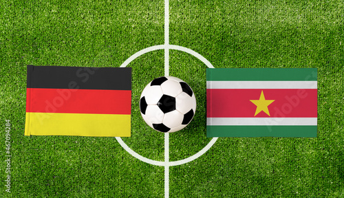 Top view soccer ball with Germany vs. Suriname flags match on green football field.