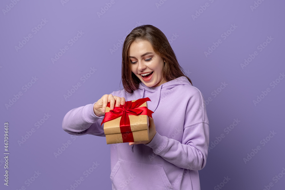 Happy young brunette woman in purple hoodie opening a present, standing over purple background. New Year Women's Day birthday holiday concept Happy young brunette woman in purple hoodie opening a present, standing over purple background. New Year Women's Day birthday holiday concept