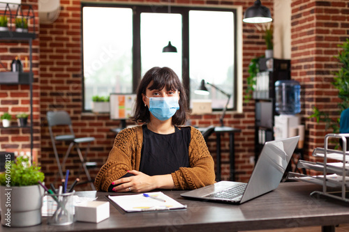 Portrait of businesswoman with medical protective face mask looking into camera working at business presentation checking financial report in startup office. Manager analyzing company strategy