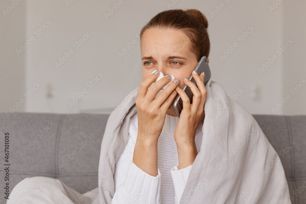 Despaired unhealthy woman with hair bun sitting on sofa wrapped in blanket talking on mobile phone, having runny nose and sneezing, has conversation with doctor. Despaired unhealthy woman with hair bun sitting on sofa wrapped in blanket talking on mobile phone, having runny nose and sneezing, has conversation with doctor.