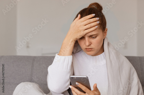 Portrait of sick unhealthy sad woman with hair bun sitting on sofa wrapped in blanket holding smart phone in hand, calling doctor, has headache, touching pain forehead.