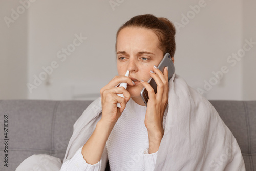 Unhealthy sad woman with hair bun sitting on sofa wrapped in blanket using nasal spray and talking with doctor  holding mobile phone in hand  posing at home.
