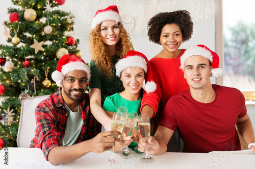 celebration and winter holidays concept - happy team in santa hats clinking glasses of non-alcoholic sparkling wine at corporate office party over christmas tree background