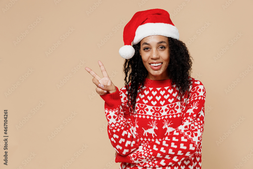 Happy young african american Santa woman in Christmas hat sweater showing victory sign isolated on pastel beige background studio portrait. Happy New Year 2022 celebration merry x-mas holiday concept. Happy young african american Santa woman in Christmas hat sweater showing victory sign isolated on pastel beige background studio portrait. Happy New Year 2022 celebration merry x-mas holiday concept.