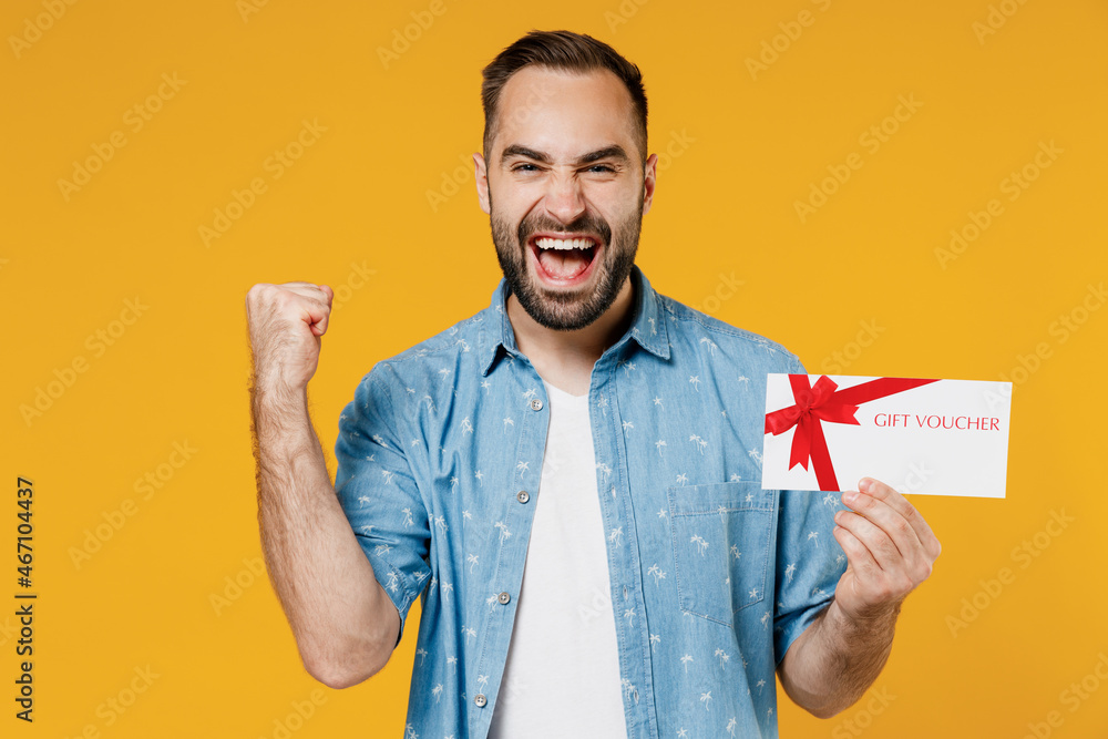 Young overjoyed fun happy caucasian man 20s wearing blue shirt white t-shirt hold gift certificate coupon voucher card for store do winner gesture isolated on plain yellow background studio portrait - obrazy, fototapety, plakaty 