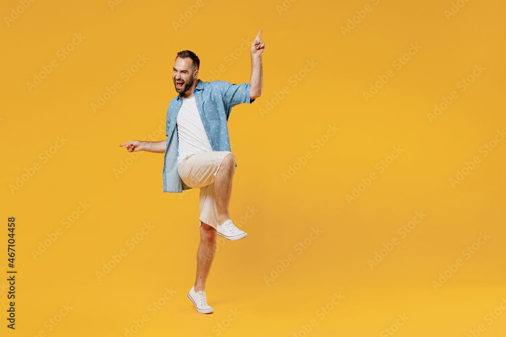 Full body young overjoyed excited caucasian man in blue shirt do winner gesture with raised up leg clench fist celebrate isolated on plain yellow background studio portrait. People lifestyle concept.