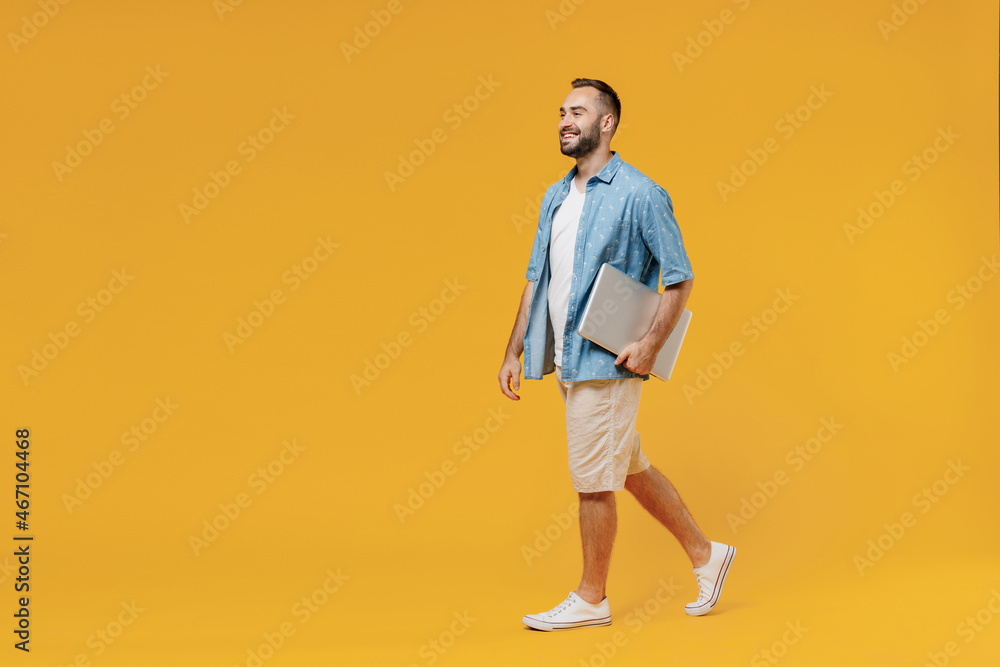 Full body side view young smiling happy fun caucasian man 20s wearing blue shirt white t-shirt use work on laptop computer hold closed pc walk go isolated on plain yellow background studio portrait Full body side view young smiling happy fun caucasian man 20s wearing blue shirt white t-shirt use work on laptop computer hold closed pc walk go isolated on plain yellow background studio portrait