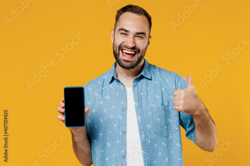 Young smiling happy man 20s in blue shirt white t-shirt hold in hand use mobile cell phone with blank screen workspace area show thump up gesture isolated on plain yellow background studio portrait.