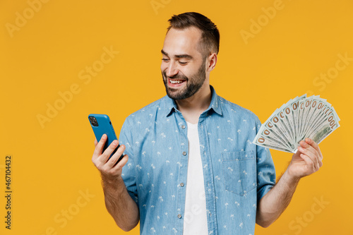 Foto Young happy caucasian man 20s wearing blue shirt holding fan of cash money in dollar banknotes mobile cell phone isolated on plain yellow background studio portrait