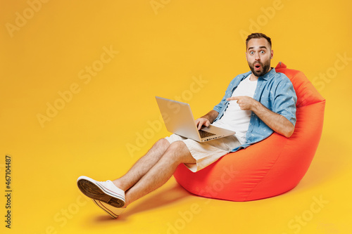 Full body young smiling fun happy man wear blue shirt white t-shirt sit in bag chair hold use work on laptop pc computer isolated on plain yellow background studio portrait. People lifestyle concept