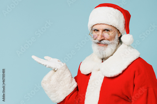 Old Santa Claus man 50s wears Christmas hat red suit clothes hold empty palm with copy space isolated on plain blue background studio. Happy New Year 2022 celebration merry ho x-mas holiday concept.