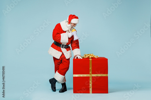 Full body old Santa Claus man 50s in Christmas hat red suit clothes stand near gift box scream shout in megaphone isolated on plain blue background studio. Happy New Year 2022 merry ho x-mas concept.