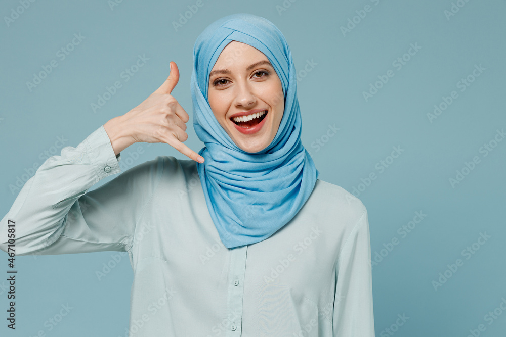 Young smiling arabian asian muslim woman in abaya hijab doing phone gesture like says call me back isolated on plain blue background studio portrait. People uae middle eastern islam religious concept. Young smiling arabian asian muslim woman in abaya hijab doing phone gesture like says call me back isolated on plain blue background studio portrait. People uae middle eastern islam religious concept.