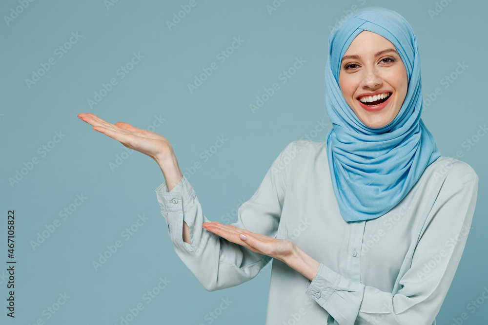 Young arabian asian muslim woman in abaya hijab point hands arms aside copy space workspace area isolated on plain blue background studio portrait. People uae middle eastern islam religious concept. - obrazy, fototapety, plakaty 