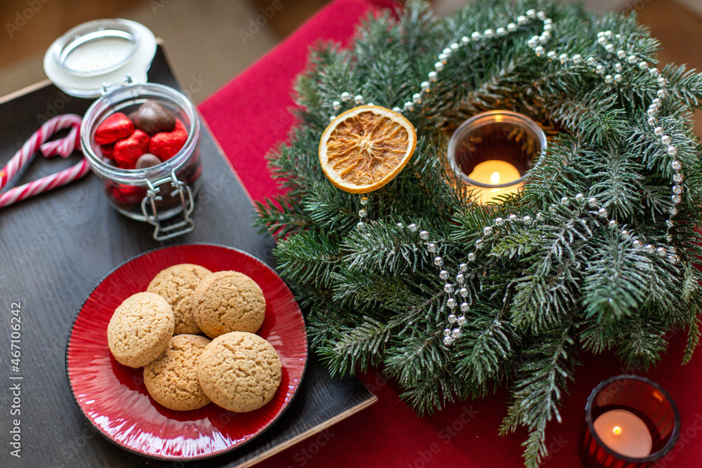bake, food and winter holidays concept - oatmeal cookies on red ceramic plate, chocolate candies and christmas wreath with burning candle at home bake, food and winter holidays concept - oatmeal cookies on red ceramic plate, chocolate candies and christmas wreath with burning candle at home