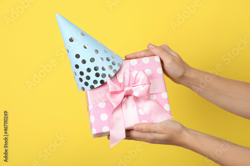 Female hands holding gift box with party hat on yellow background
