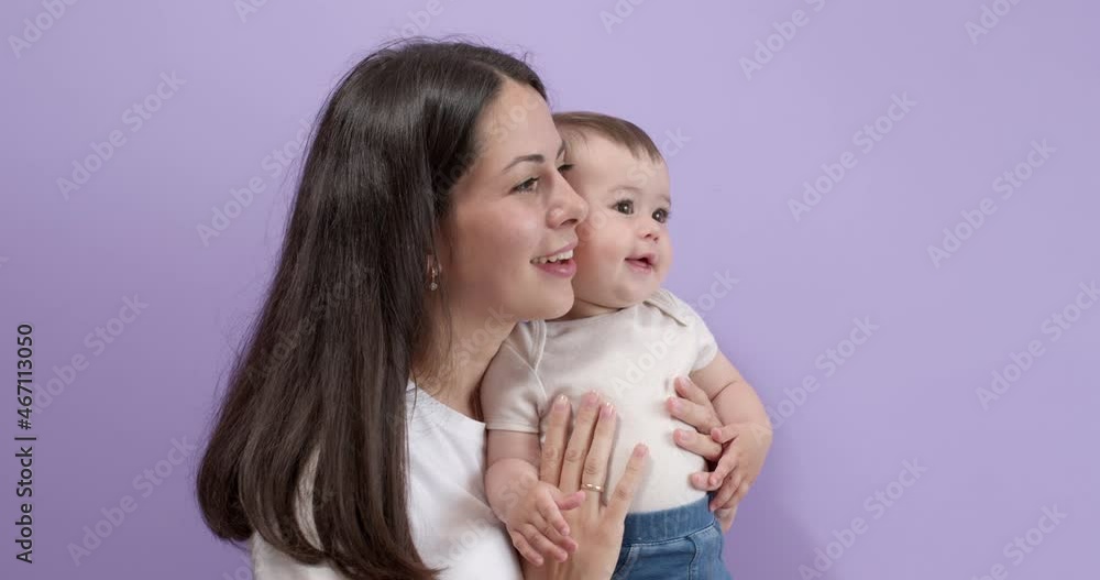 Beautiful Caucasian Brunette Female Holding Little Child Girl Daughter Carefully In Hands Looking At Side Talking, Smiling. Woman In Casual Wear Posing With Toddler Child, Isolated On Purple Beautiful Caucasian Brunette Female Holding Little Child Girl Daughter Carefully In Hands Looking At Side Talking, Smiling. Woman In Casual Wear Posing With Toddler Child, Isolated On Purple