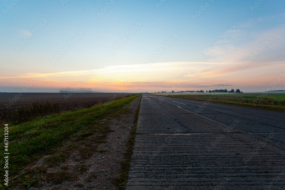 Fototapeta premium An empty asphalt road through the fields and forest in a thick fog at sunrise