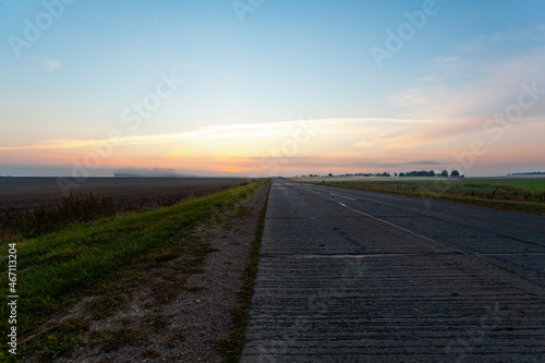 An empty asphalt road through the fields and forest in a thick fog at sunrise