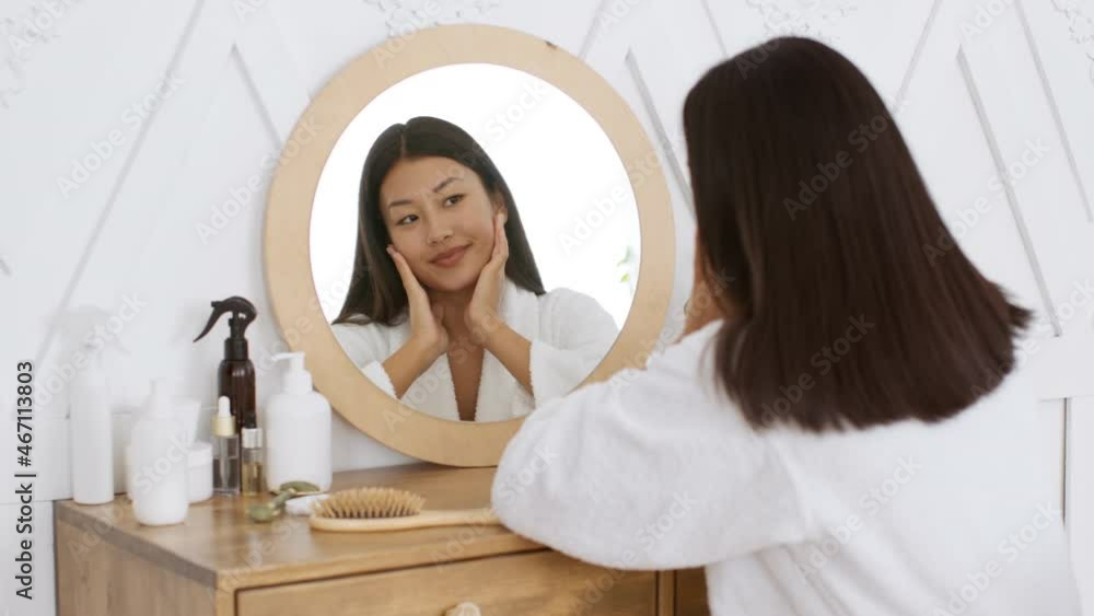 Young attractive korean woman looking at mirror, smiling to herself, enjoying her youth and beauty, sitting at bathroom Young attractive korean woman looking at mirror, smiling to herself, enjoying her youth and beauty, sitting at bathroom