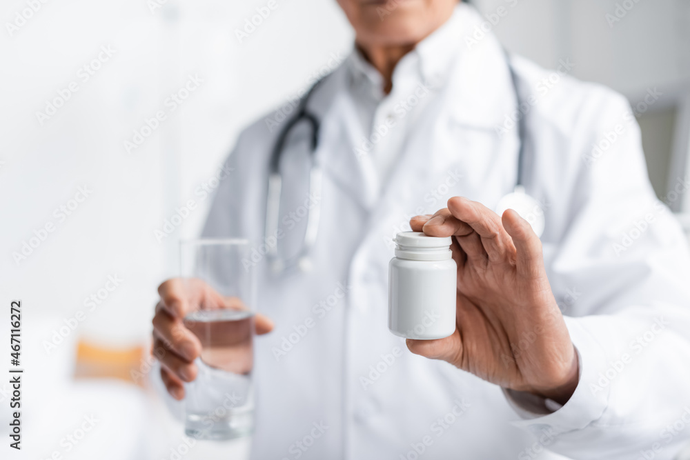 Cropped view of blurred doctor holding jar with pills and glass of water in clinic Cropped view of blurred doctor holding jar with pills and glass of water in clinic