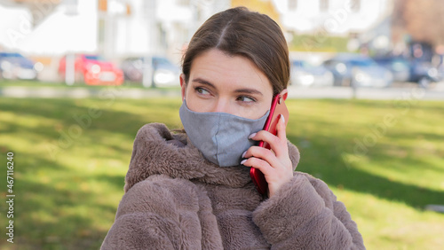 Close up of young pretty Caucasian woman standing in city in face mask and talking on smartphone. Portrait of beautiful female calling and chatting on cellphone outdoors in quarantine. Leisure concept