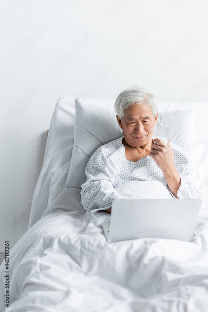 Senior asian patient holding glass of water and looking at laptop on bed in clinic Senior asian patient holding glass of water and looking at laptop on bed in clinic