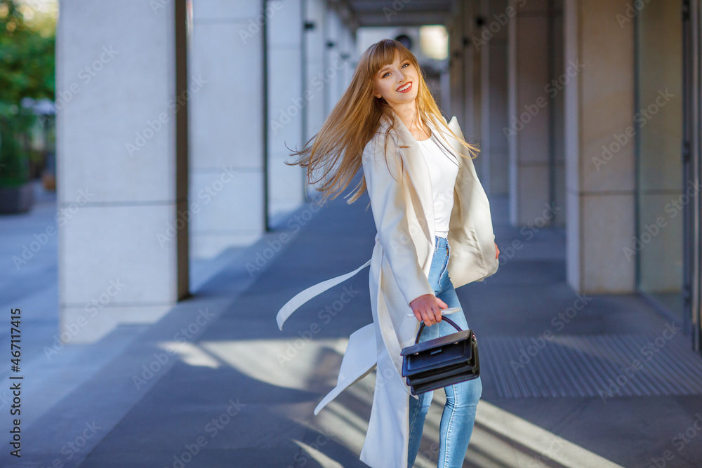 Happy young adult woman smiling with teeth smile, walking outdoors on city street, fluttering hair. Attractive girl weared in autumn clothes. White coat, long hair. Blurred details