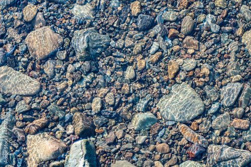 Sun glare on surface water and smooth stones at bottom stream, background. Transparent water in river with rocky bottom, fast flow
