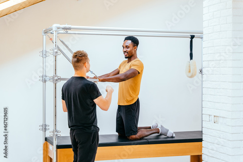 Serious male trainer helping a african european man in pulling stretch bands sitting on pilates training machine. Sportive man at the gym doing pilates training with his trainer.