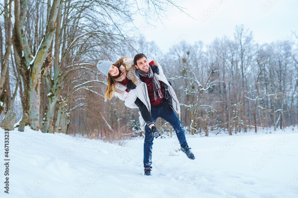 Happy young man carry his smiling pretty woman on his back at winter park. Boyfriend giving piggyback ride on shoulder to his girlfriend in winter holiday.